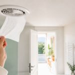 Main tenant une feuille de papier toilette devant une bouche d’extraction de VMC dans une salle de bains lumineuse